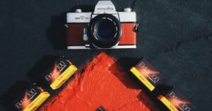 A Minolta SRT 101 camera with a red textured grip sits above four boxes of Kodak Ektar 100 film arranged on a dark surface with a red painted shape.
