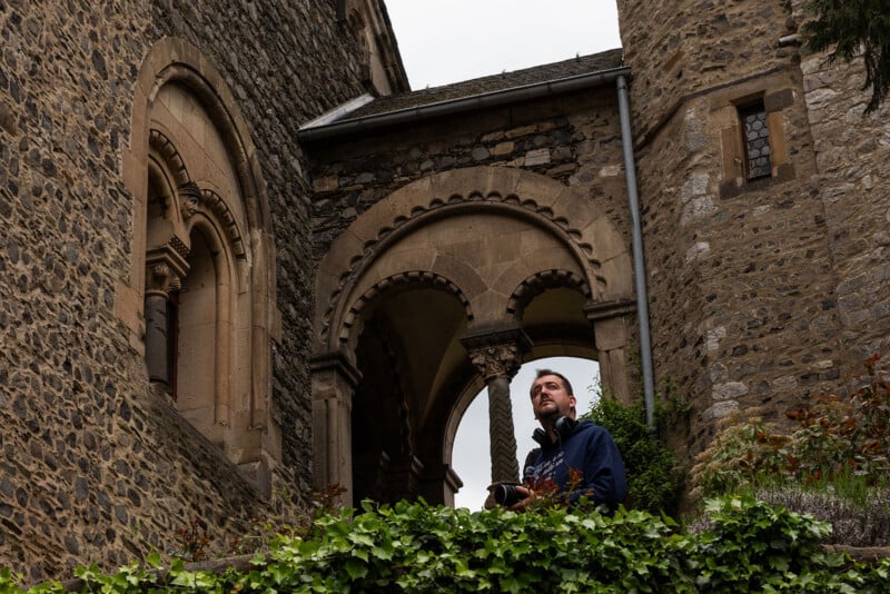 A man holding a camera stands in front of a stone archway connecting two sections of an old, ivy-covered stone building.