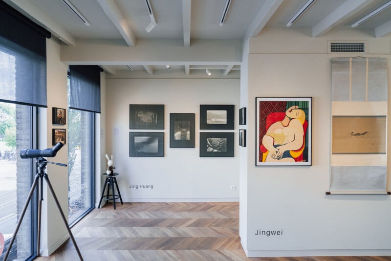 An art gallery interior features black-and-white photographs on a wall labeled "Jing Huang," a Picasso print, and a hanging scroll labeled "Jingwei," with a tripod-mounted telescope in the foreground.