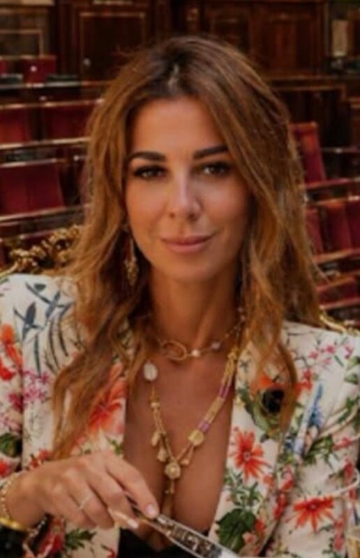 A woman with long, wavy brown hair wears a floral-patterned blazer and layered gold necklaces while holding a piece of cutlery.