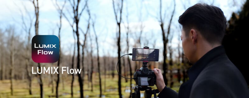 A person stands behind a camera mounted on a tripod, with a smartphone attached to the top of the camera displaying a live feed, next to the text "LUMIX Flow".