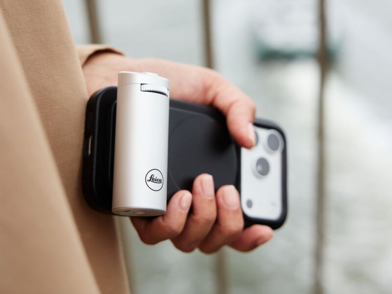 A person holds a smartphone with a silver, cylindrical Leica-branded accessory attached to the back.