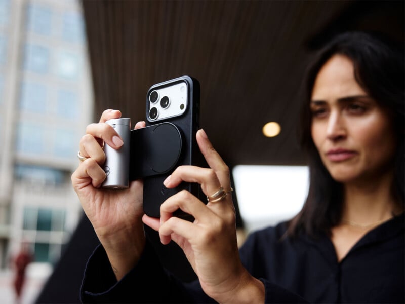A woman holds a smartphone equipped with a silver, cylindrical camera grip attachment while looking toward the side.