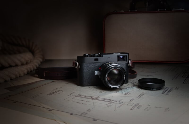 A black Leica camera with a lens sits on a technical blueprint next to a lens cap, a leather strap, and a brown case.