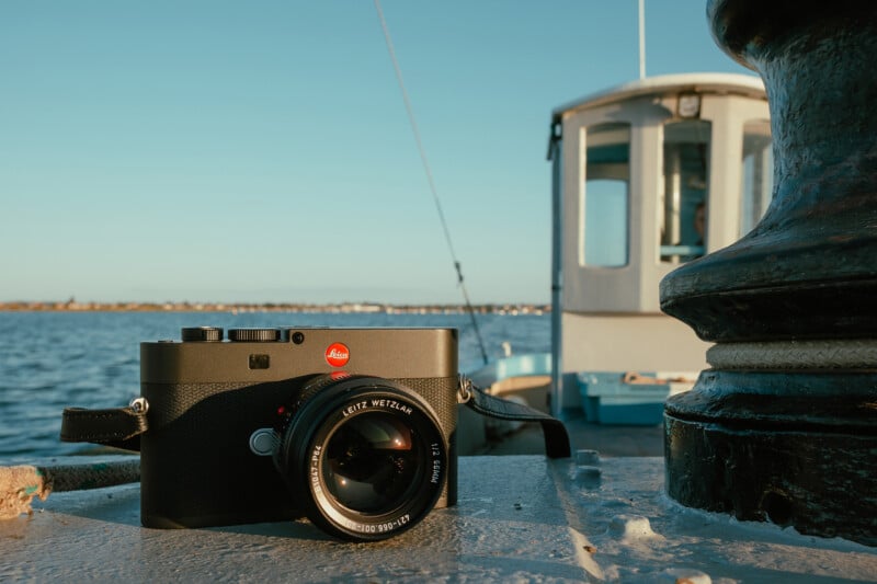 A black Leica camera with a Leitz Wetzlar lens sits on a metal surface next to a large bollard, with a boat and water in the background.