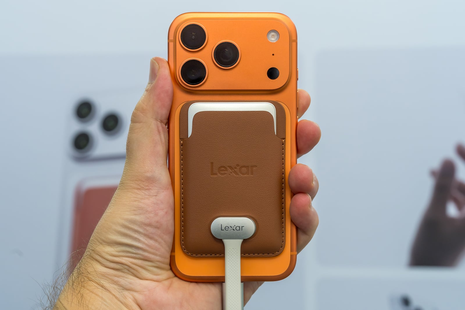 A hand holds an orange smartphone with a brown leather Lexar-branded magnetic wallet attached to the back, connected by a white cable.