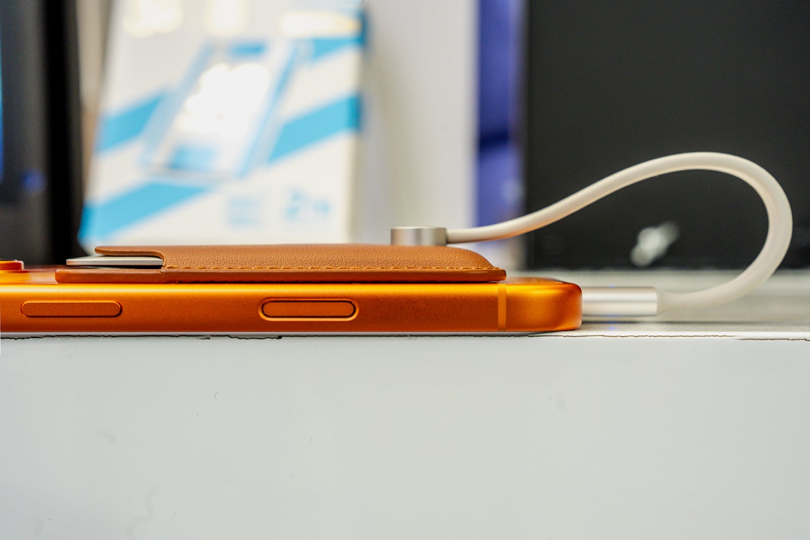 A side view of an orange smartphone with a brown leather card holder attached to the back, connected to a white charging cable.
