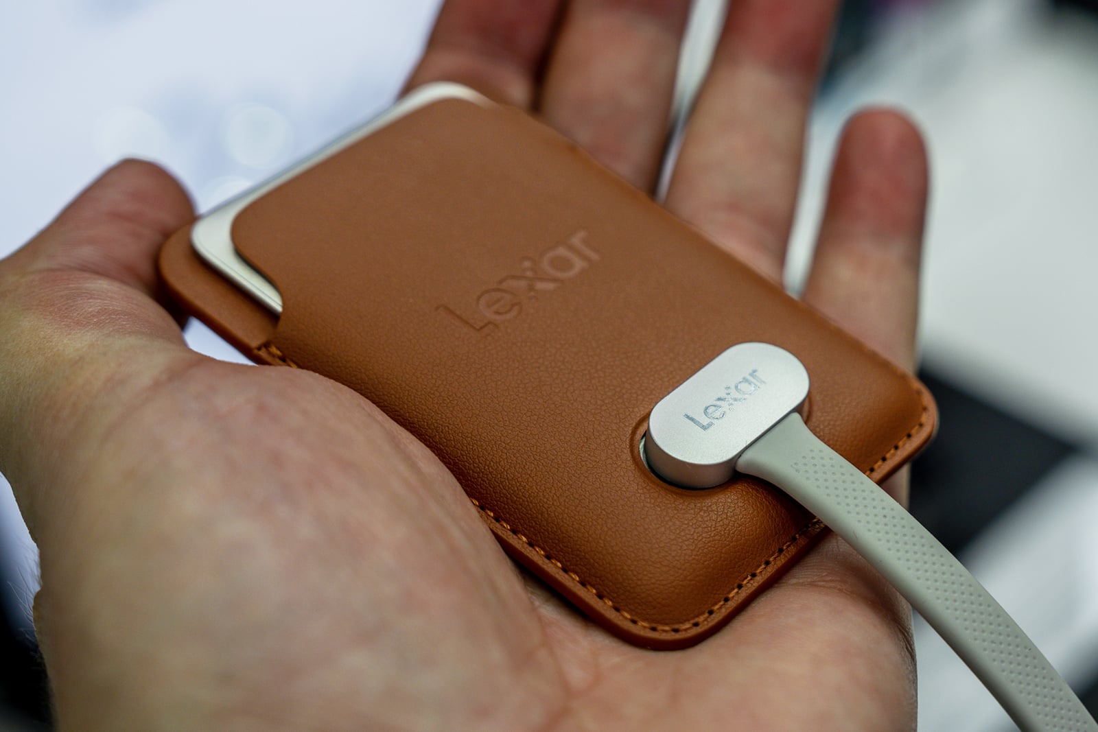 A hand holds a brown leather Lexar-branded portable SSD with a silver USB-C cable plugged into the side.