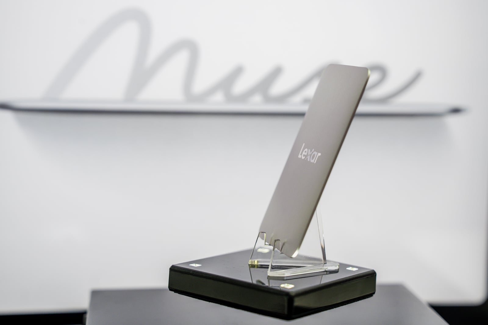 A slim, silver Lexar portable solid-state drive stands upright on a clear plastic display stand atop a black base, with the word "Muse" written in cursive on the wall behind it.