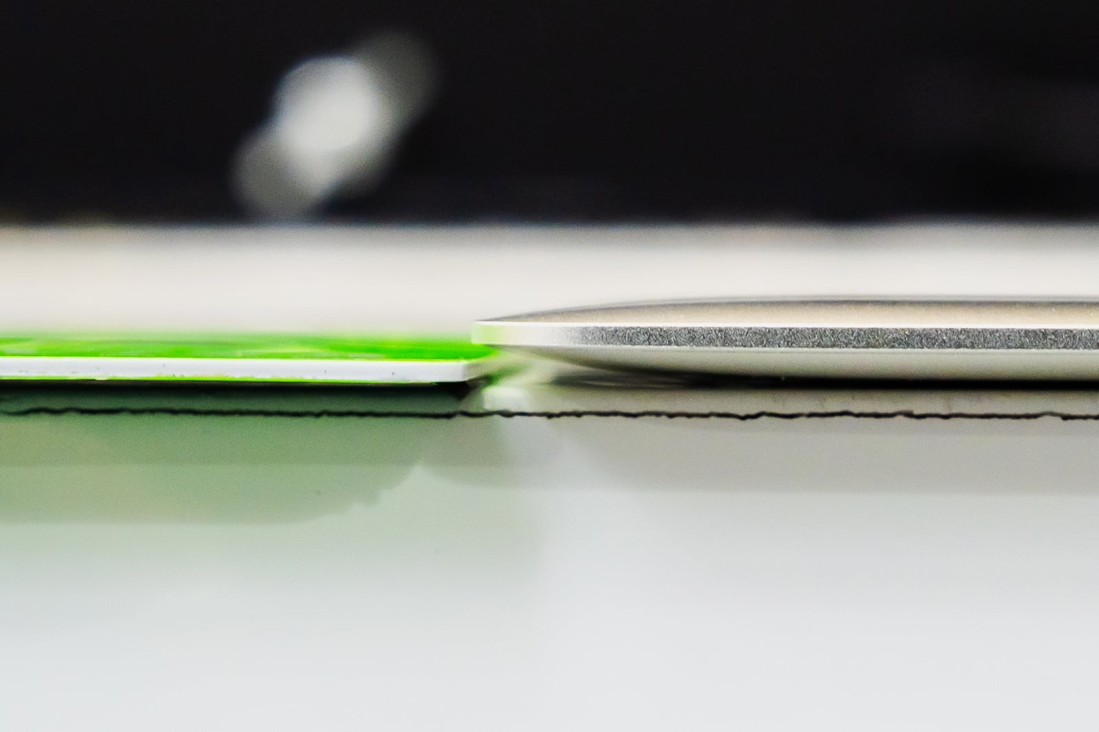 A side-by-side close-up shows a thin, flat green card next to a thicker, rounded metallic object resting on a white surface.
