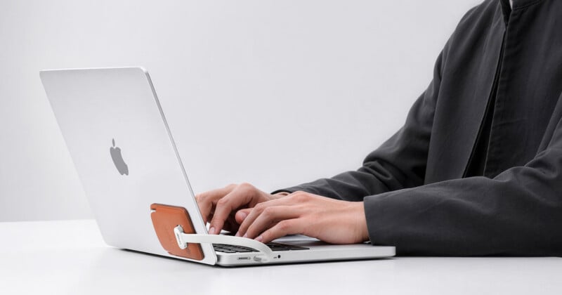 A person in a dark jacket types on a silver laptop with a small, tan leather accessory attached to the back of the screen by a white cable.