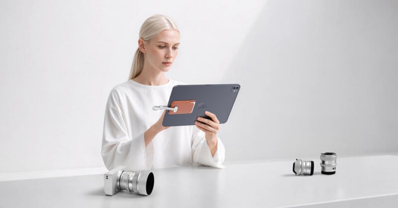 A woman with blonde hair wearing a white shirt holds a tablet with a brown magnetic accessory attached to the back, while sitting at a table with camera lenses.