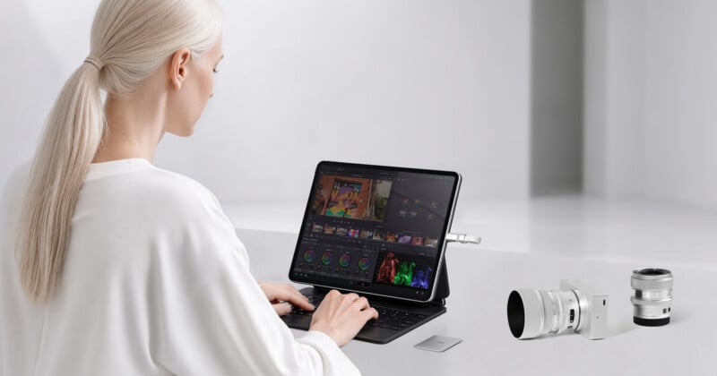 A person with blonde hair in a ponytail sits at a desk, using a tablet with a keyboard cover to edit video, with a camera, a lens, and a small memory card placed on the table nearby.