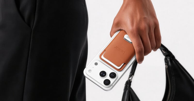 A person holds a white smartphone with a brown leather magnetic card wallet attached to the back, alongside a black handbag.