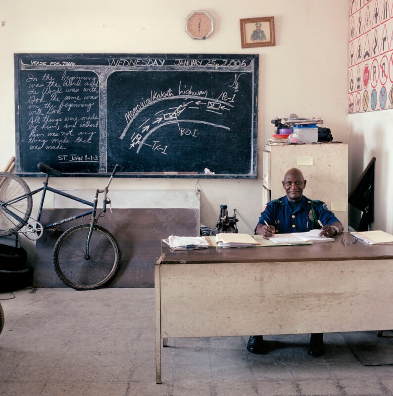 A police officer sits at a desk writing in a logbook in front of a chalkboard displaying a traffic diagram and a Bible verse.