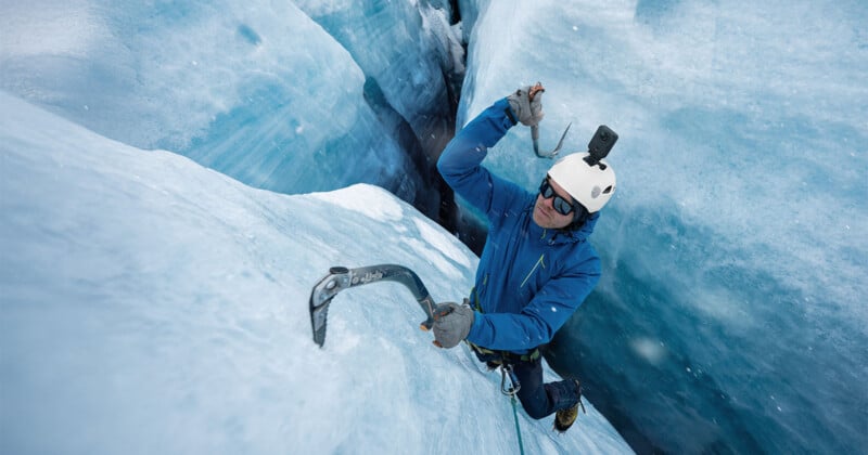 A climber in a blue jacket and white helmet with a mounted camera scales a steep, blue ice wall using two ice axes.