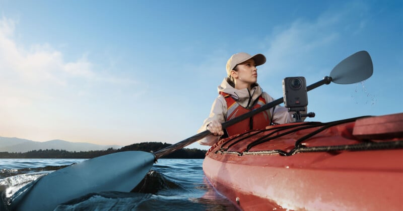 A person wearing a cap and life vest paddles a red kayak on a lake under a clear blue sky, with a small camera mounted to the deck.