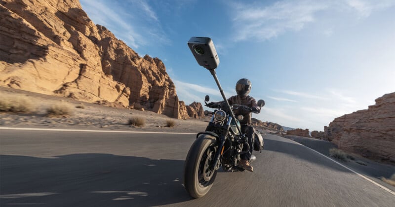 A person wearing a helmet and leather jacket rides a motorcycle on a desert road, with a tall camera mount extending upward from the handlebars.