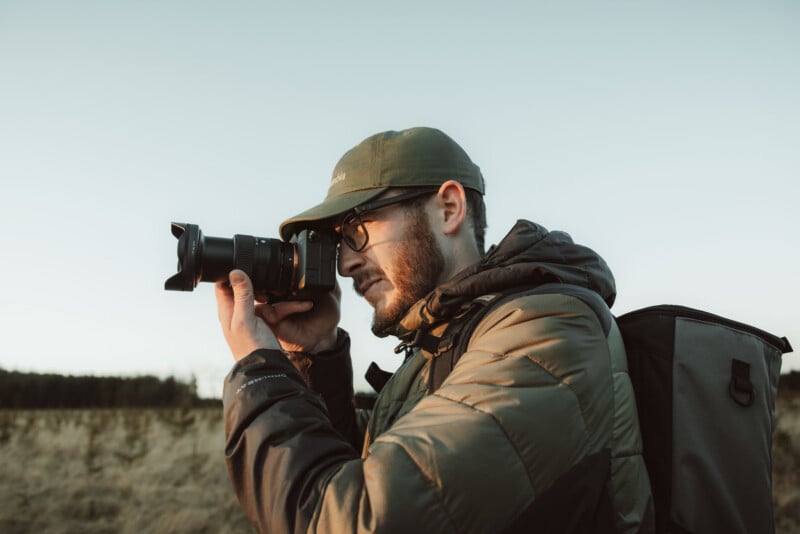 A man with a beard, glasses, and a green baseball cap holds a camera to his eye while wearing a backpack in a field.