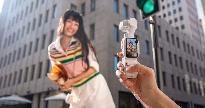 A person holds a small, white handheld gimbal camera in the foreground, displaying a live view of a smiling woman holding a paper bag on a city street.
