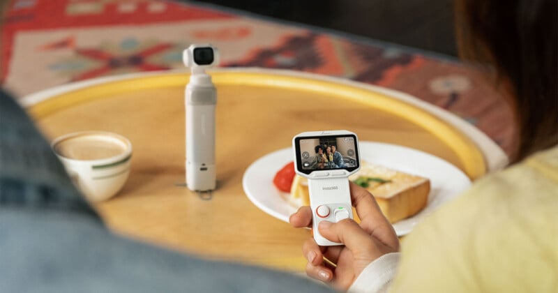 A person holds a white handheld camera with a screen displaying a live feed of three people, while a separate white gimbal camera stands on a wooden table next to a cup of coffee and a plate of food.