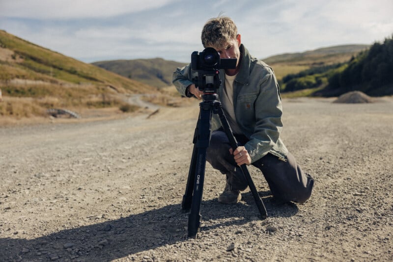 A person kneels on a gravel path, adjusting a camera mounted on a tripod while looking through the viewfinder.