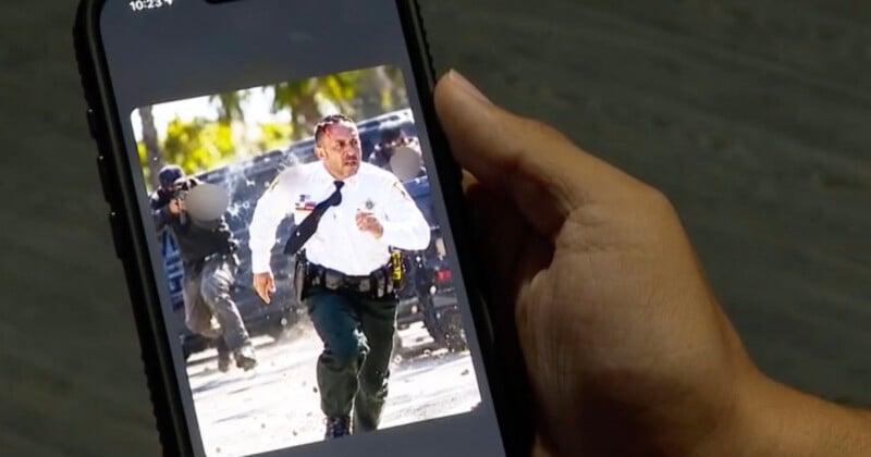 A hand holds a smartphone displaying a photo of a law enforcement officer running away from a scene with a tactical team in the background.