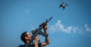 A man aims a rifle upward at a small drone flying against a clear blue sky.