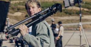 A young man with blonde hair carries a tripod on his shoulder while standing on a film set with crew members in the background.