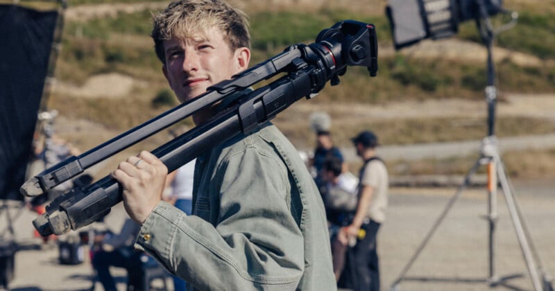 A young man with blonde hair carries a tripod on his shoulder while standing on a film set with crew members in the background.