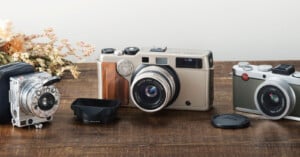 Three vintage cameras and a lens hood sit on a wooden table next to a small bouquet of dried flowers.