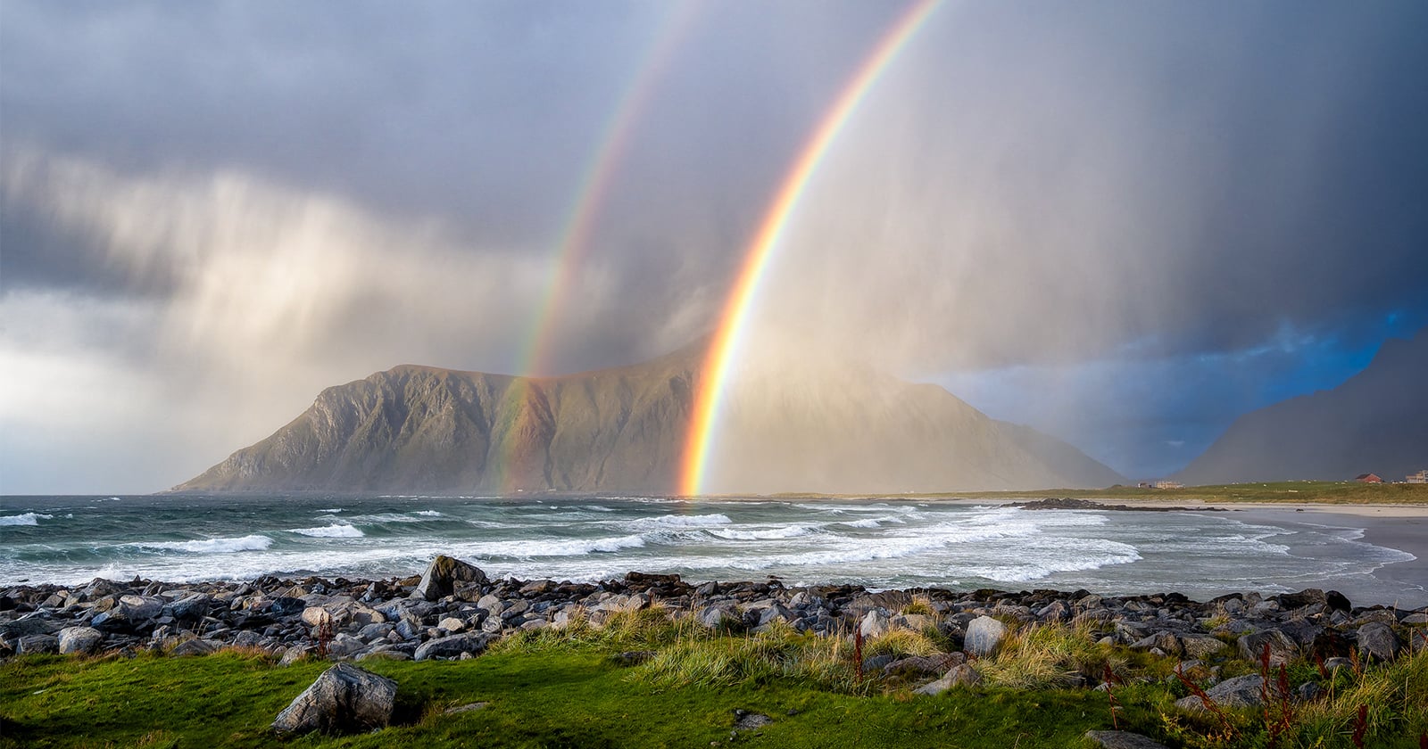 A double rainbow arches over a rocky coastline and a large mountain under a stormy, cloudy sky.