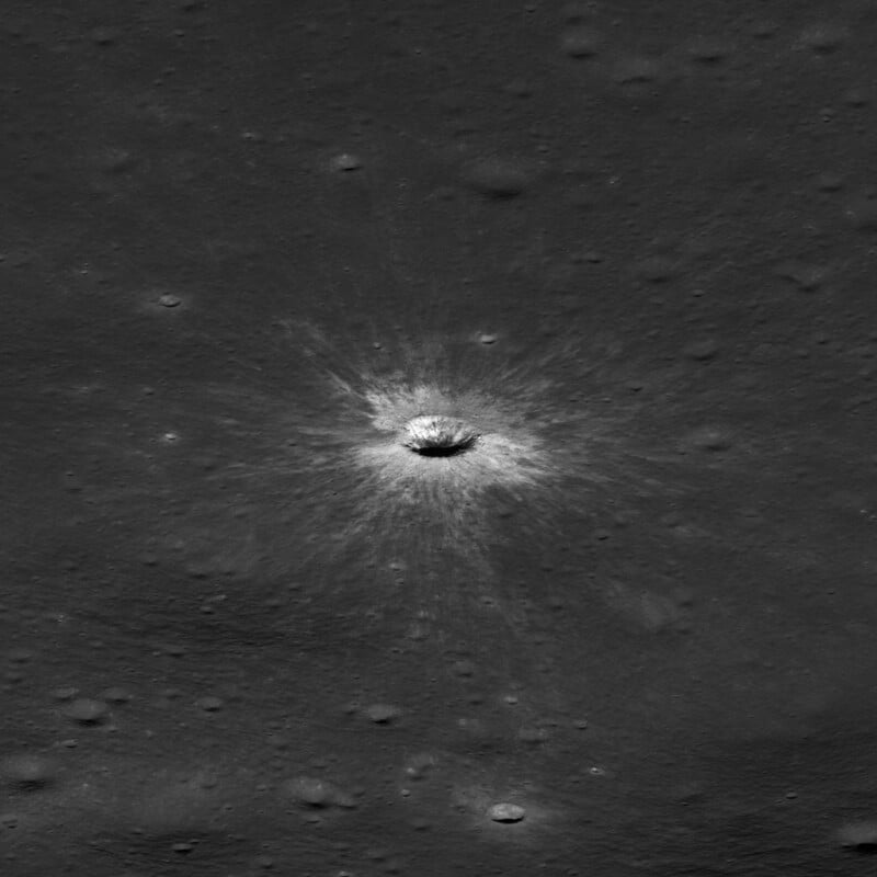 A small, bright impact crater with a prominent radial ray system sits in the center of a dark, smooth lunar surface.