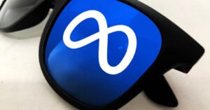 A pair of black sunglasses with the white Meta infinity logo reflected on the blue-tinted lens.