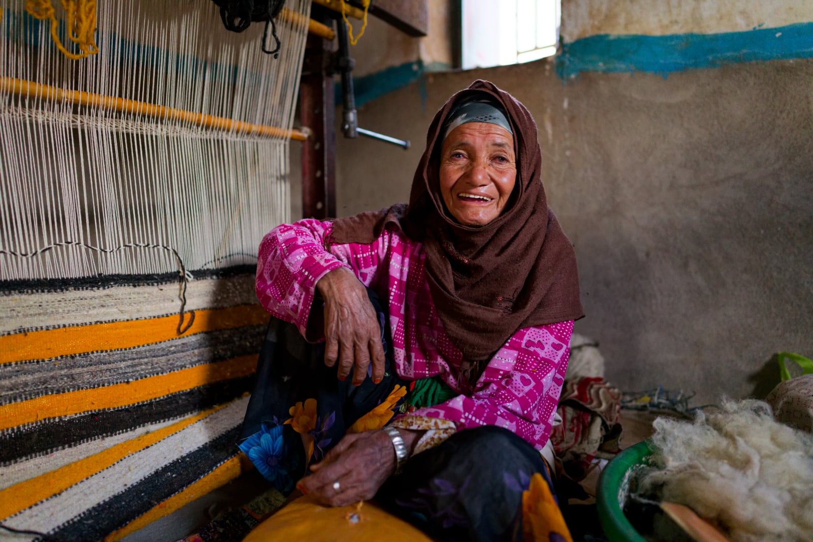 An elderly woman wearing a brown headscarf and a pink patterned shirt sits smiling in front of a traditional wooden loom.
