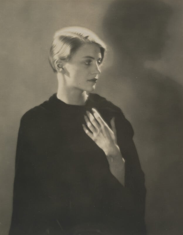 A black-and-white portrait of a person with short blonde hair, wearing a dark garment and holding one hand to their chest, set against a soft, shadowed background.