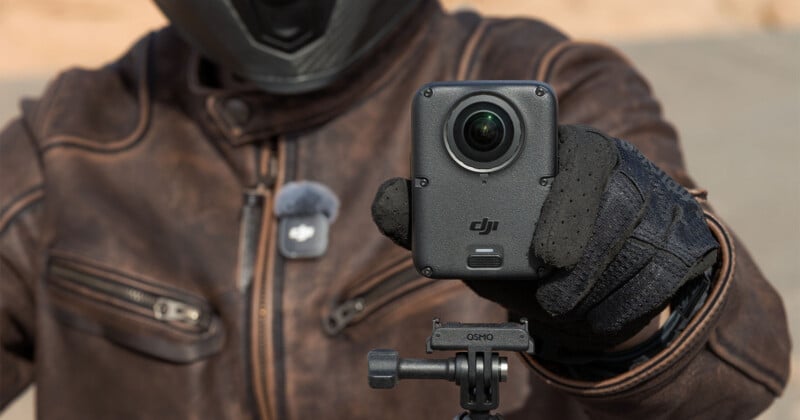 A person wearing a leather jacket and motorcycle gloves holds a black DJI Osmo action camera mounted on a tripod.