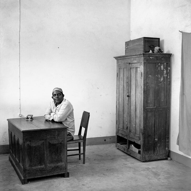 A man wearing a patterned cap and light-colored shirt sits at a wooden desk in a sparsely furnished room with a tall wooden cabinet.