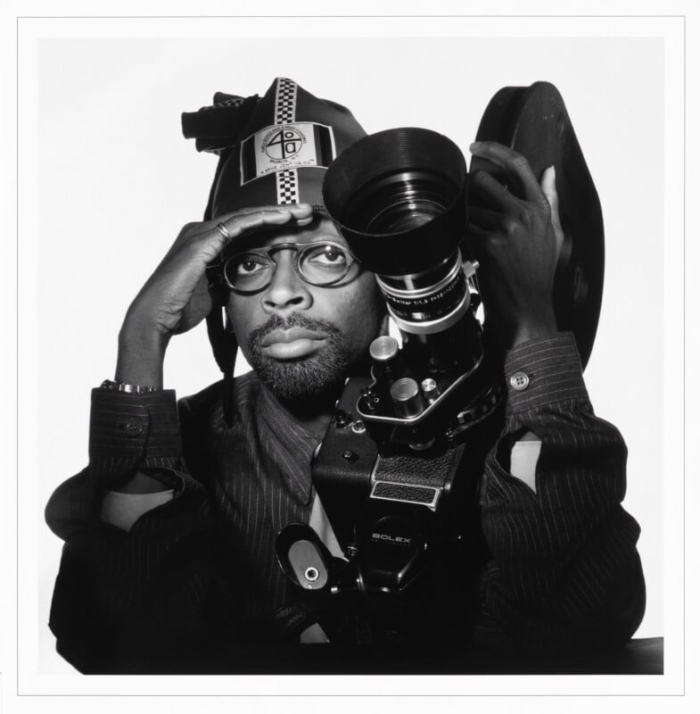 Spike Lee poses in a black-and-white portrait, wearing a branded hat and glasses while holding a Bolex movie camera.
