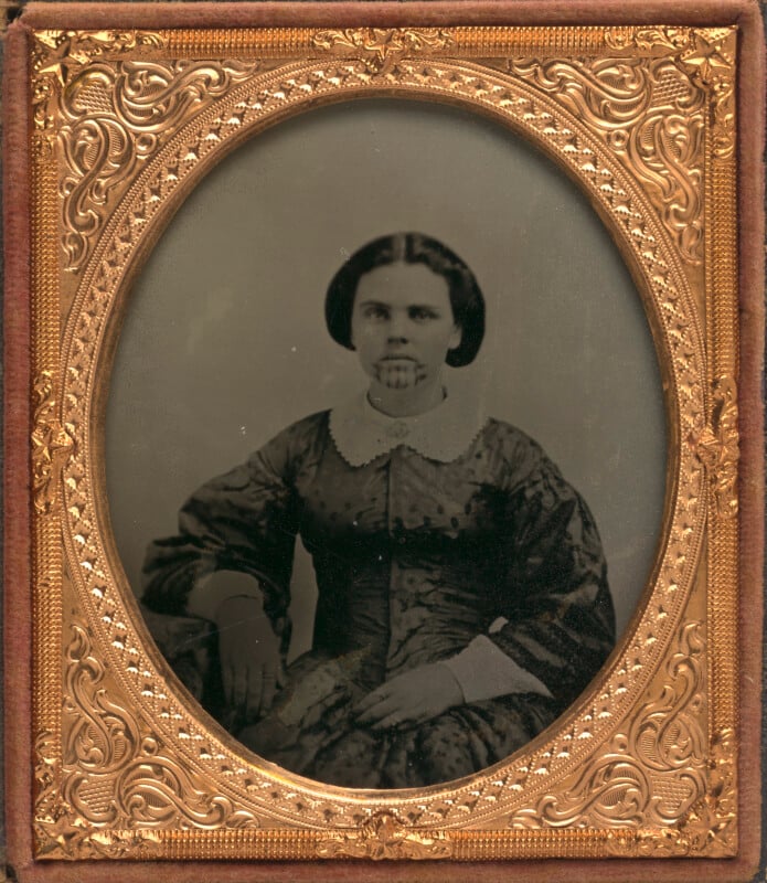 A daguerreotype portrait of a woman with a chin tattoo, wearing a dark patterned dress with a white collar, set in an ornate gold oval frame.