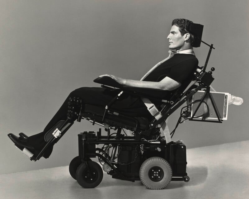 Christopher Reeve sits in profile in a motorized wheelchair, wearing a black shirt and supported by a headrest and ventilator.