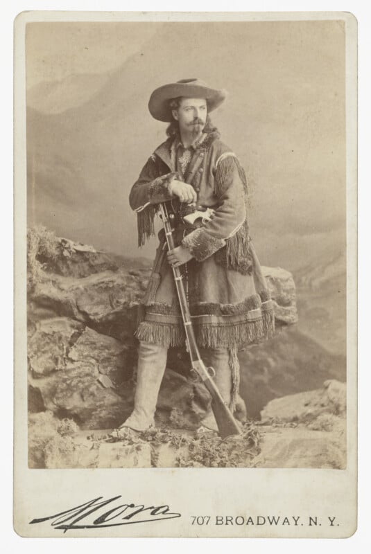 A studio portrait of William "Buffalo Bill" Cody standing on a rock formation, wearing a fringed buckskin coat and holding a rifle.