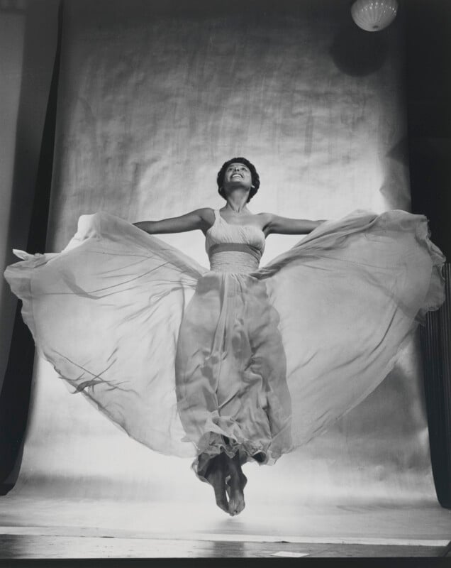 A woman with short dark hair jumps in the air, wearing a flowing, light-colored gown that spreads out around her against a plain backdrop.
