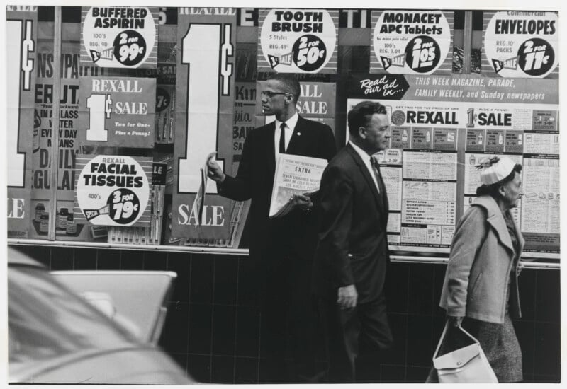 Malcolm X walks past a Rexall store window filled with "1¢ Sale" advertisements, holding a newspaper with a headline about a police shooting.