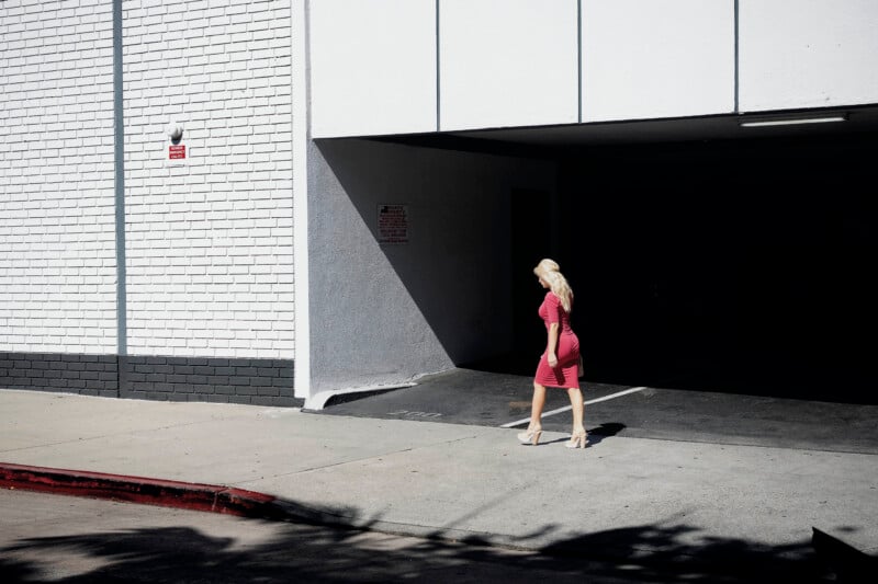 A blonde woman in a pink dress and high heels walks past the entrance of a parking garage on a sunny day.
