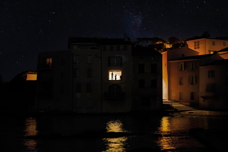 A couple stands in a brightly lit window of a dark building overlooking a body of water under a starry night sky.