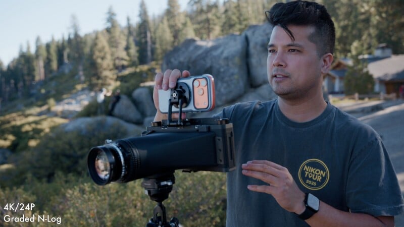 A man wearing a Nikon Tour 2025 t-shirt stands outdoors, holding a smartphone mounted to a large, specialized camera lens on a tripod.
