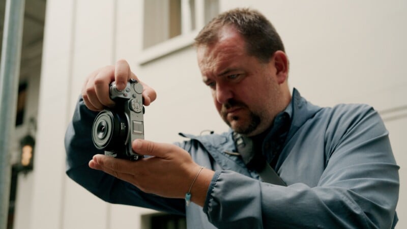 A man with a goatee wearing a blue jacket holds a silver digital camera with both hands while looking down at its screen.
