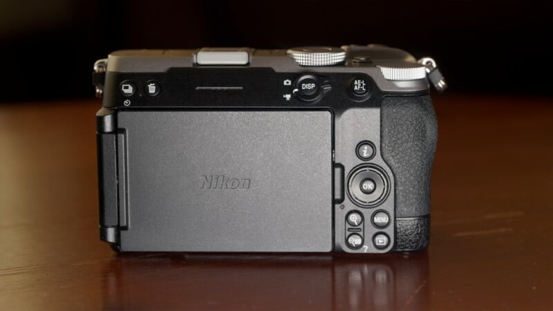 The back of a black Nikon mirrorless camera sits on a wooden surface, showing its LCD screen, control buttons, and dials.