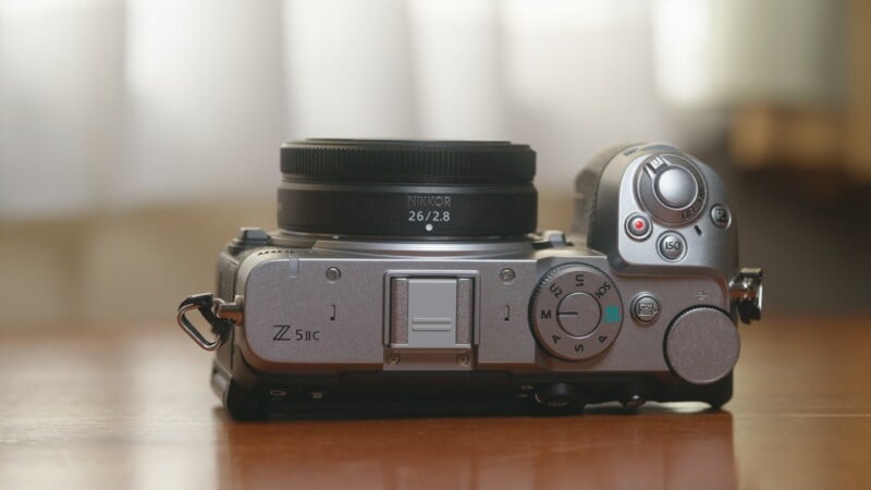 A silver Nikon Z fc mirrorless camera with a Nikkor 26mm f/2.8 lens sits on a wooden surface.